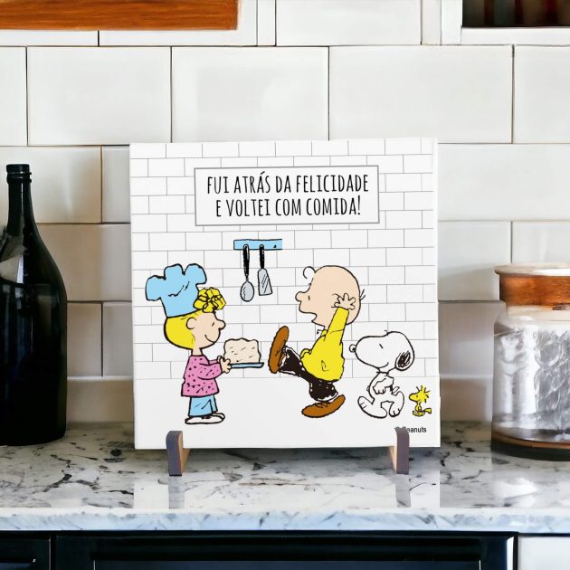 Na foto um azulejo decorativo criativo e engraçado com desenho do Snoopy com um boné e o woodstock em uma cozinha com louças na pia. No azulejo tem a frase enquanto houver vida haverá louça na pia.