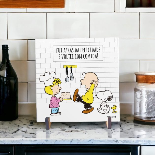 Na foto um azulejo decorativo criativo e engraçado com desenho do Snoopy com um boné e o woodstock em uma cozinha com louças na pia. No azulejo tem a frase enquanto houver vida haverá louça na pia.